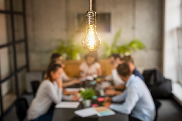 a light bulb is in focus above a blurred background showing a meeting between several people