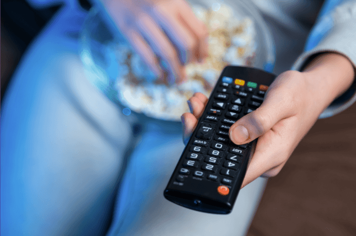 a person holding a tv remote and a bowl of popcorn sits on the couch