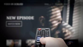A hand holding a TV remote points to the TV screen that shows a new episode of a streaming series