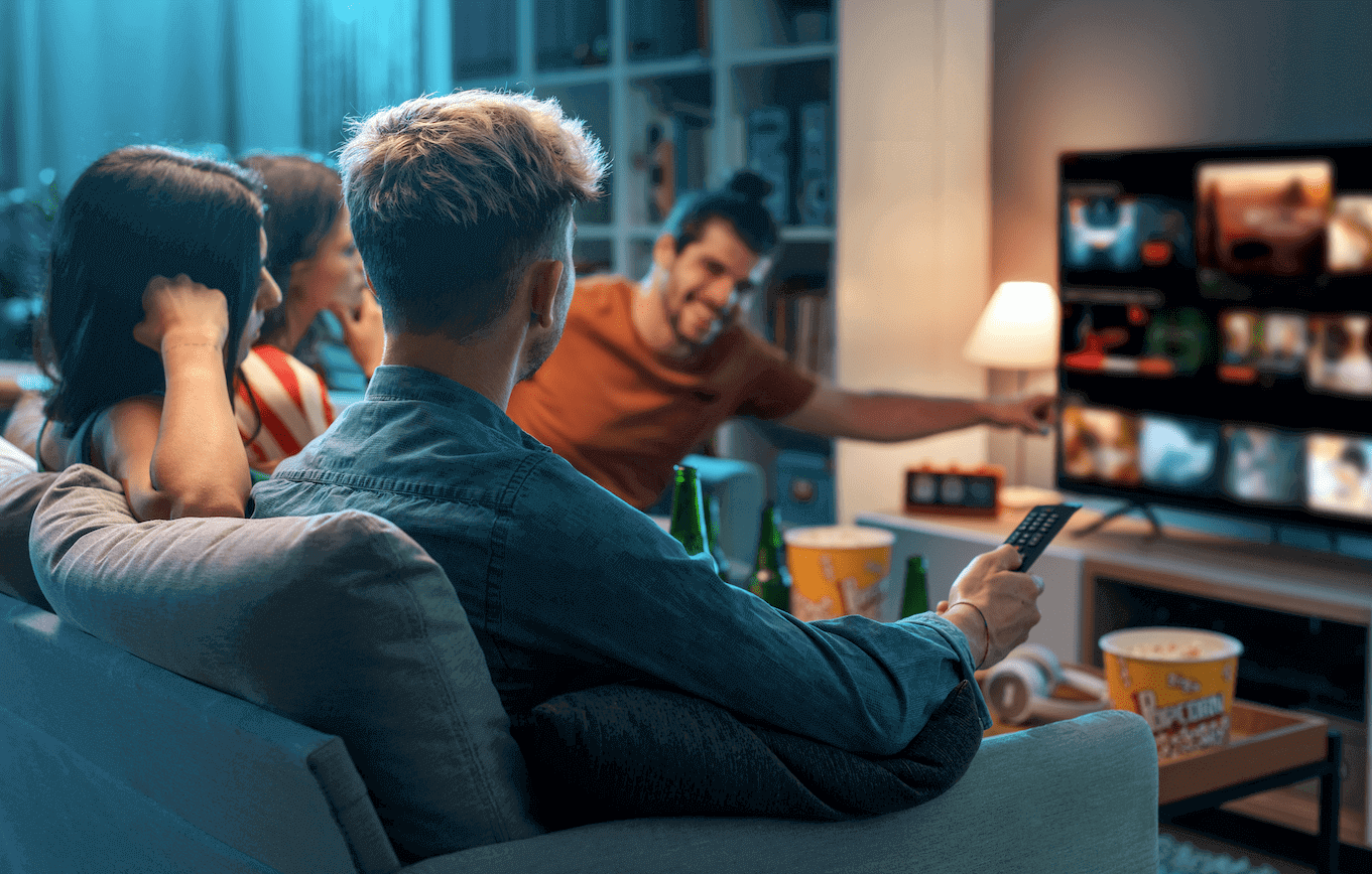four people on the couch are browsing a streaming service and one points to the TV screen