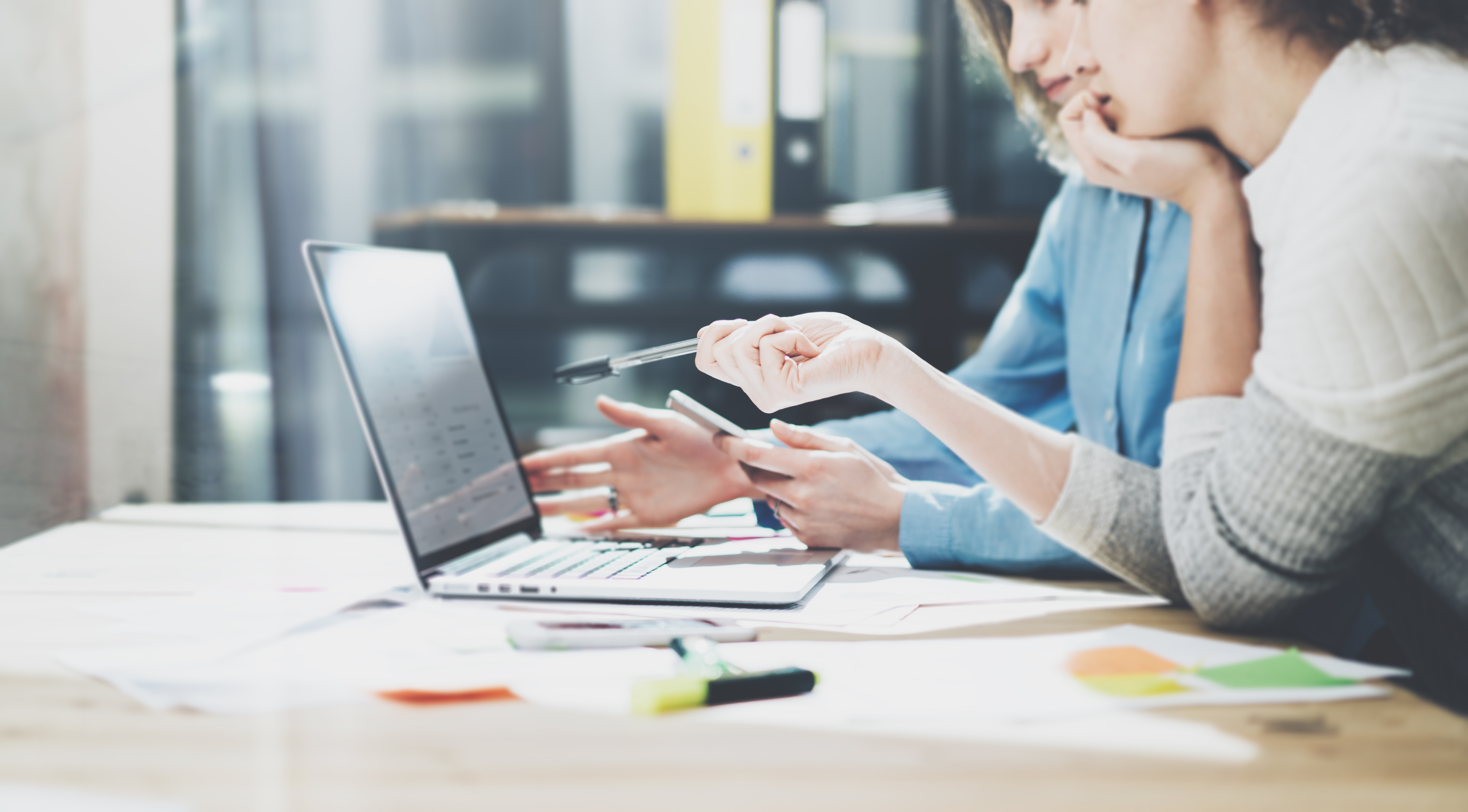 two women work side by side pointing to a laptop surrounded by papers